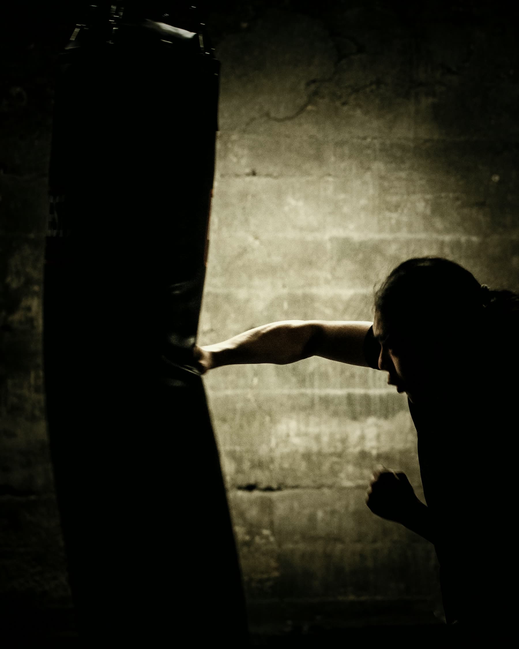 A person working a heavy bag in a dark room at night, lit by a single warm light, face in shadow
