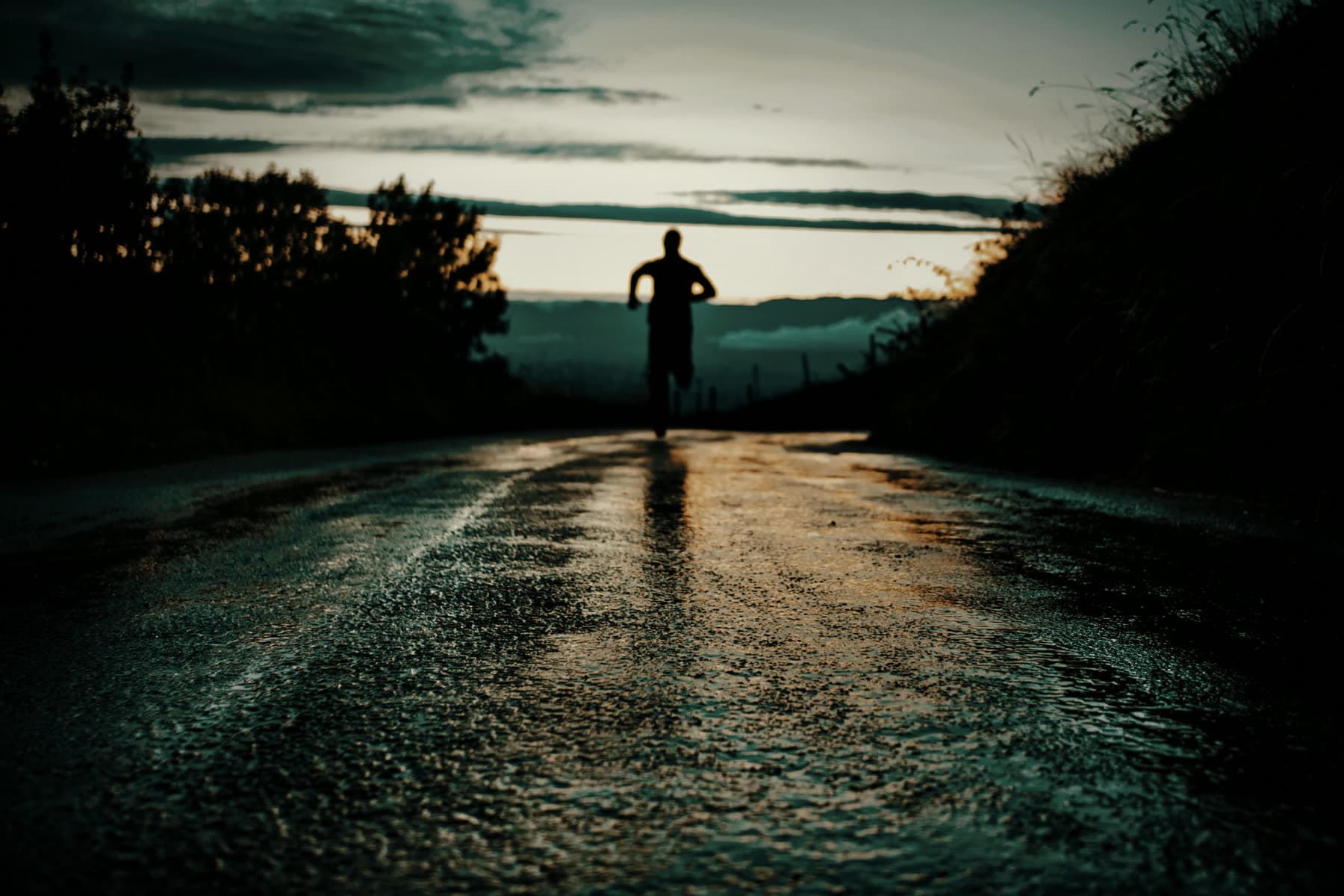 A lone runner in silhouette on a wet road at dawn, warm light reflecting off the asphalt