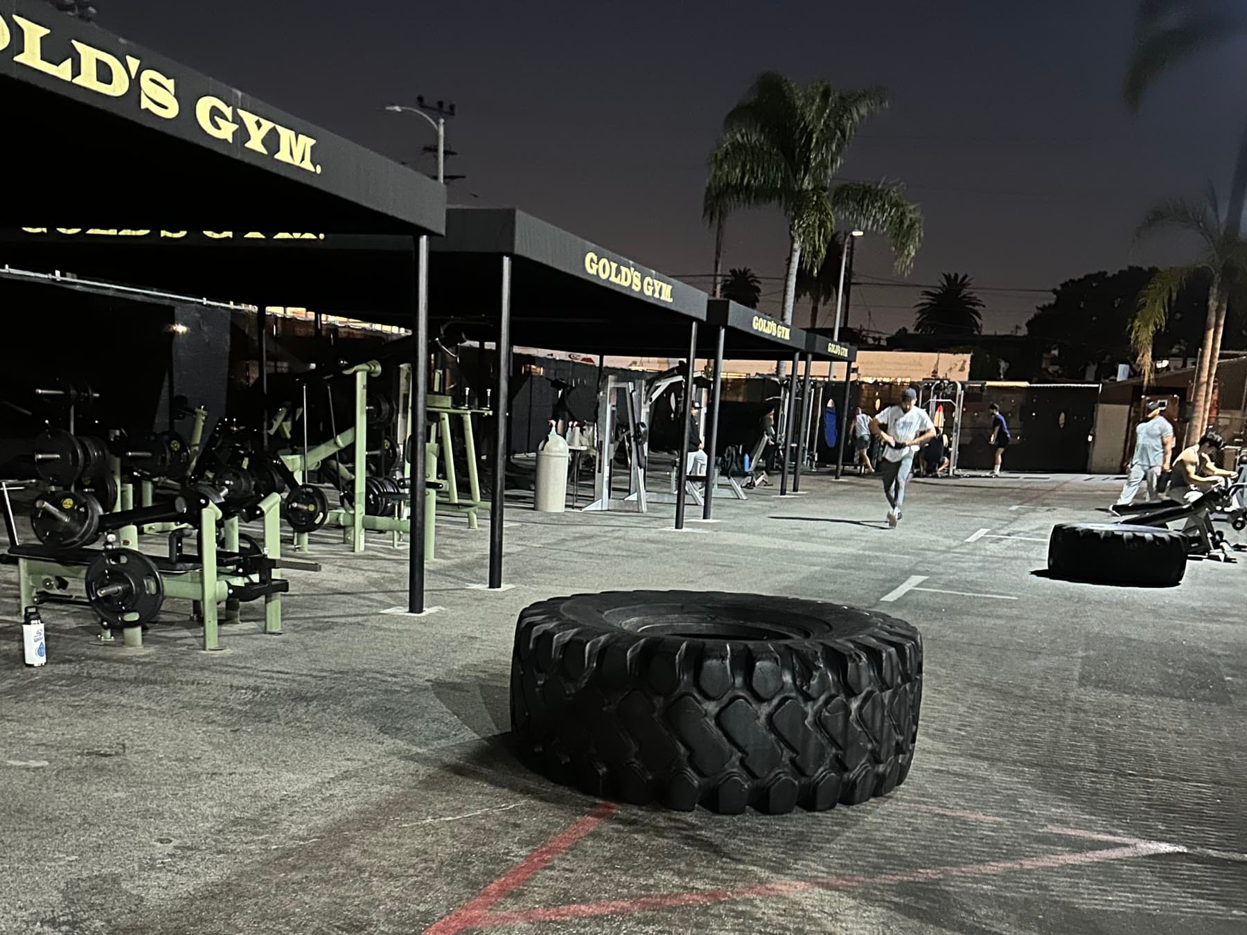 The outdoor training pit at Gold's Gym Venice at night, a giant flip tire resting on the lot beneath the canopies