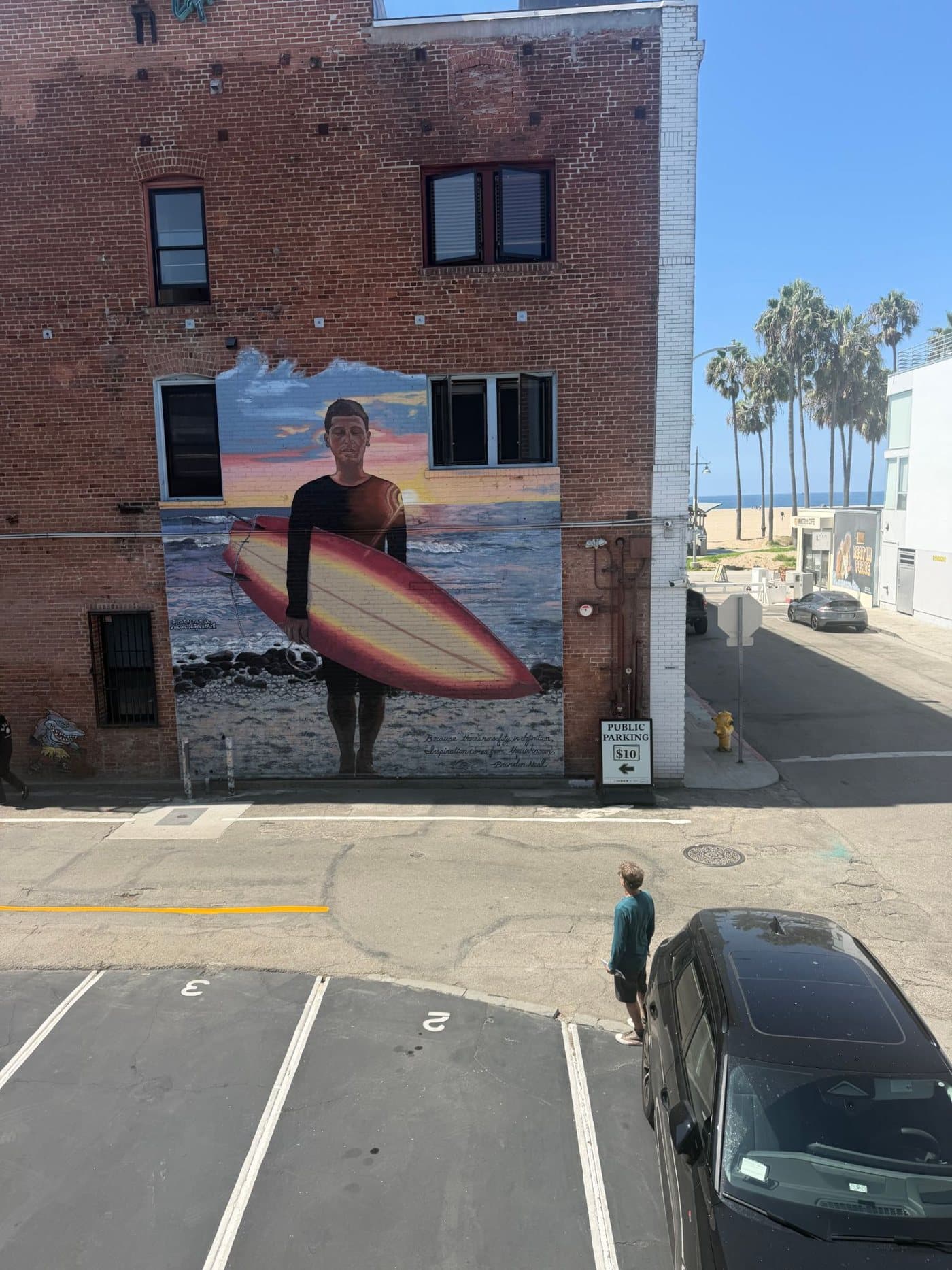 From Marc's balcony: Brad Neal stands in the street looking up at the mural of his son Brandon holding a surfboard, palm trees and the beach beyond
